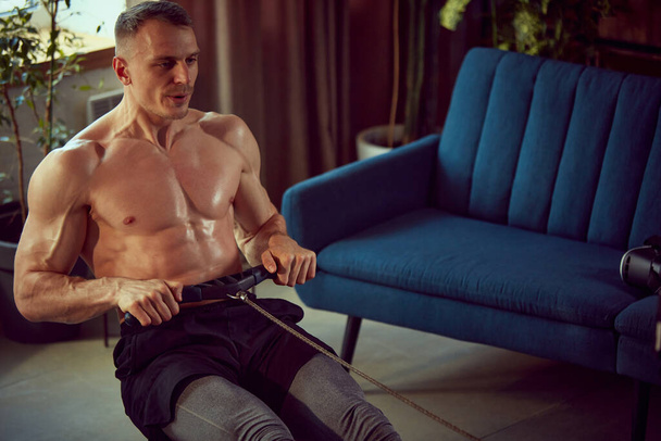 Handsome young man with relief, muscular body training shirtless, doing exercises with stationary rowing machine. Concept of sportive lifestyle, body and health care, fitness, health - Foto, imagen