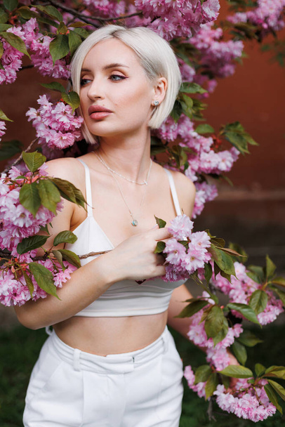 Mujer sexy con el pelo blanco corto en la luz del sol sobre un fondo beige y una rama de sakura floreciente. - Foto, imagen
