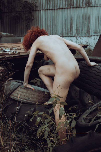 A completely naked red-haired woman poses among old tires in a scrap yard and used agricultural machinery - Foto, Bild