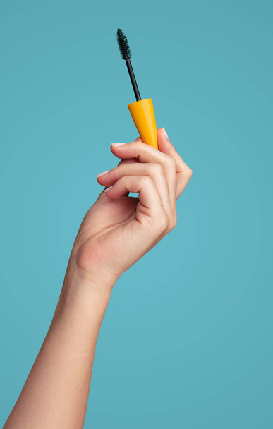 Crop anonymous female hand showing mascara brush with yellow cap against blue background - Φωτογραφία, εικόνα
