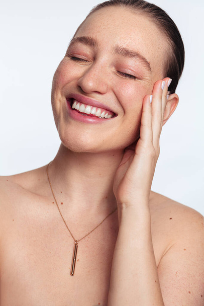 Confident happy young female with freckles and bare shoulders smiling brightly with closed eyes while touching face against white background in studio - Foto, Bild