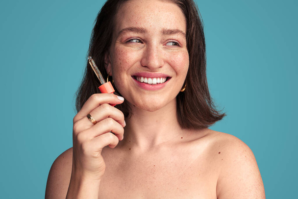 Smiling young female model with dark hair and freckled skin holding dropper with serum for skincare treatment against turquoise background - Foto, Bild