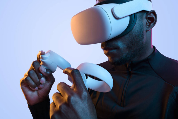 Crop serious young bearded African American man in VR goggles playing video game using controllers in neon studio - Foto, Imagen