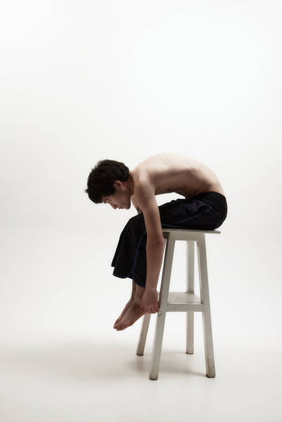 Concentration on inner self. Image of young man posing shirtless in black trousers on high chair against white studio background. Concept of male body aesthetics, style, fashion, health, mens beauty - 写真・画像