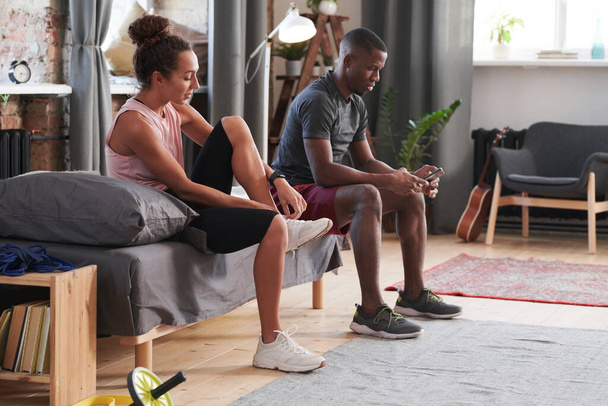 Ethnically diverse man and woman sitting on sofa in living room putting on trainers to go out for jogging in morning - Valokuva, kuva
