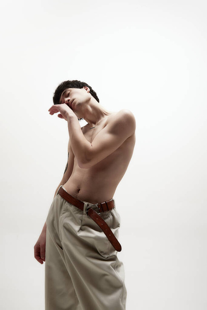 Self-care. Portrait of young handsome man with fit body, posing shirtless in beige pants against white studio background. Concept of male body aesthetics, style, fashion, health, mens beauty - 写真・画像