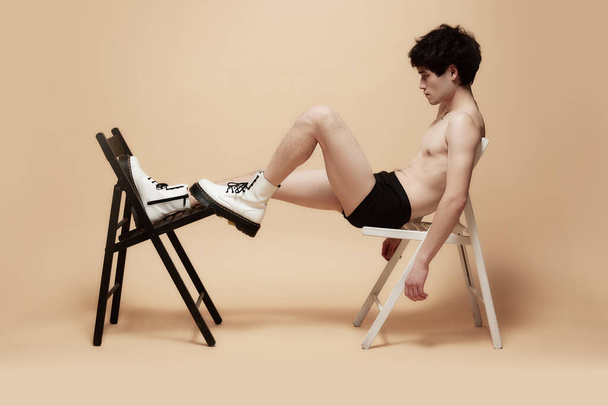 Young asian guy in underwear and stylish boots, posing shirtless on chair against light brown studio background. Concept of male body aesthetics, style, fashion, health, mens beauty - Foto, Bild