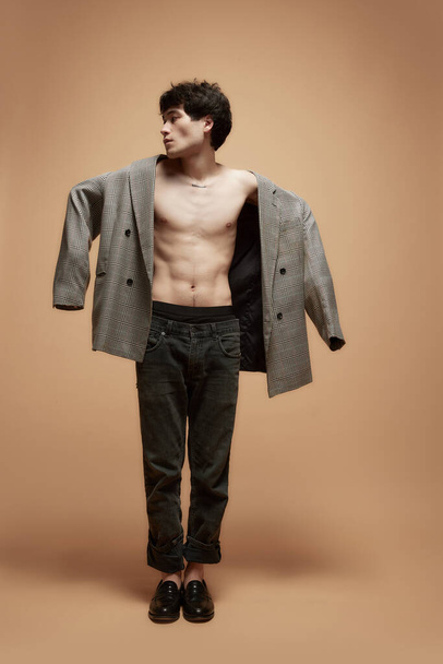 Full-length portrait of young asian man posing shirtless in stylish grey jacket and pants against light brown studio background. Male body aesthetics, style, health, mens beauty and fashion concept - Foto, imagen