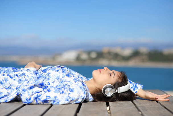 Woman relaxing listening to music with headphone lying in wooden platform on the beach - Foto, imagen