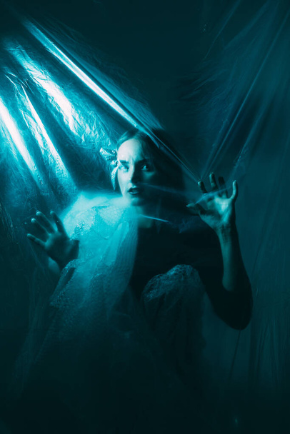 Horror fear. Night terror. Paranormal mystery. Blue color light defocused scared terrified woman wrapped in polyethylene film trapped behind plastic transparent wall on dark. - Foto, Imagem