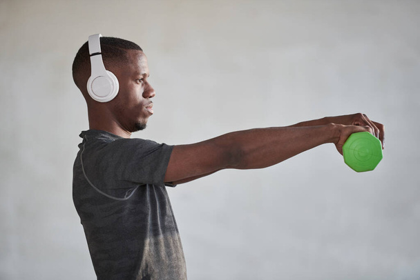 Οριζόντια μεσαία πλαϊνή εικόνα του νεαρού μαύρου άνδρα φορώντας λευκά ακουστικά ακούγοντας μουσική και κάνοντας άσκηση με αλτήρα - Φωτογραφία, εικόνα