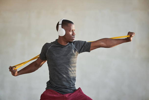 Horizontal medium shot of young Black man doing exercise with resistance band wearing headphones looking away - Foto, Bild