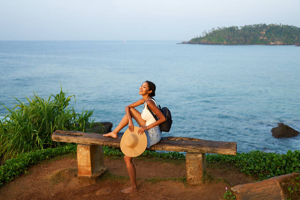 Multiethnic woman in straw hat enjoys tropical vacation sitting on rocky cliff with sea view. Black female with backpack sightseeing on scenic location. Girl tanning on island on ocean sunset. - Foto, imagen