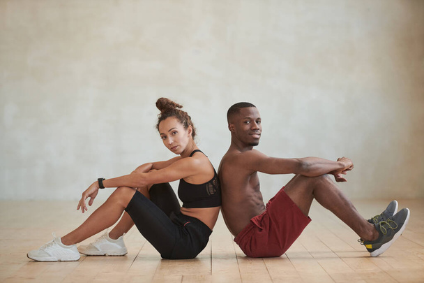 Studio shot of sporty young Black man and Caucasian woman sitting back to back in loft studio room looking at camera - Fotó, kép