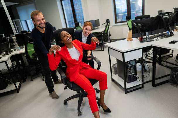 Caucasian red-haired woman, bearded caucasian man rolled African American young woman on office chair. Colleagues have fun at work - Photo, Image