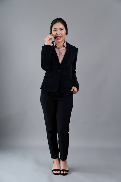 Attractive full body asian female call center operator with happy face advertises job opportunity on empty space, wearing formal suit and headset on customizable isolated background. Enthusiastic - Zdjęcie, obraz