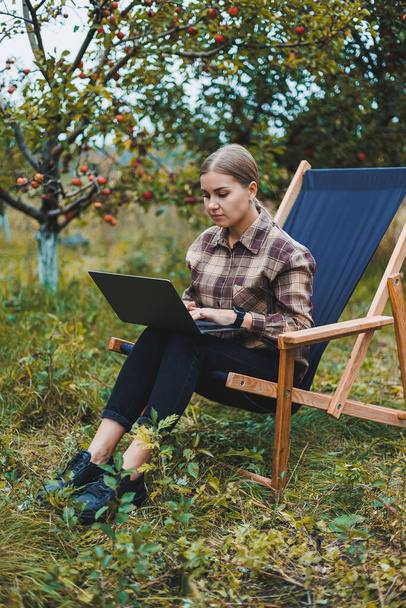 A cute young female freelancer is working in the garden outdoors while sitting in a comfortable garden chair. Remote work on a portable laptop - Foto, immagini