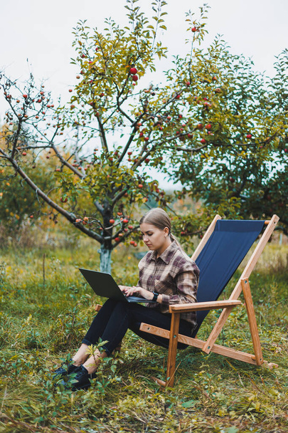 Söpö nuori naisfreelancer työskentelee puutarhassa ulkona istuessaan mukavassa puutarhatuolissa. Etätyö kannettavalla kannettavalla tietokoneella - Valokuva, kuva