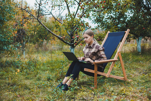 Eine süße junge Freiberuflerin arbeitet im Garten, während sie in einem bequemen Gartenstuhl sitzt. Fernbedienung auf einem tragbaren Laptop - Foto, Bild