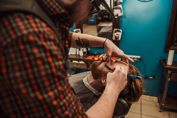 A professional barber cuts his beard to a young hipster man. Beard trim at the barbershop. Stylish beard cut. - Fotó, kép