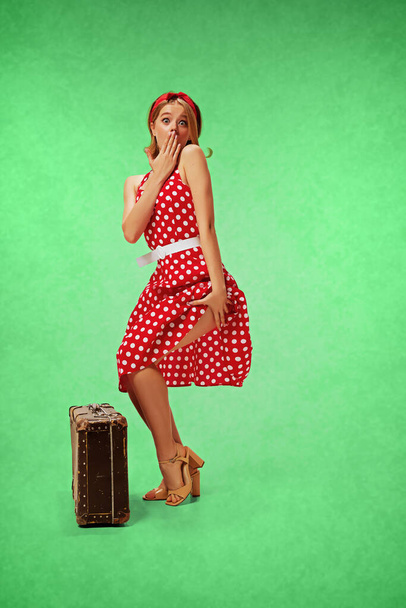 Coquette travelling. Pretty, cute young girl in red stylish dress posing with vintage suitcases against green studio background. Concept of retro fashion, beauty, 50s, 60s. Pin-up style - Φωτογραφία, εικόνα