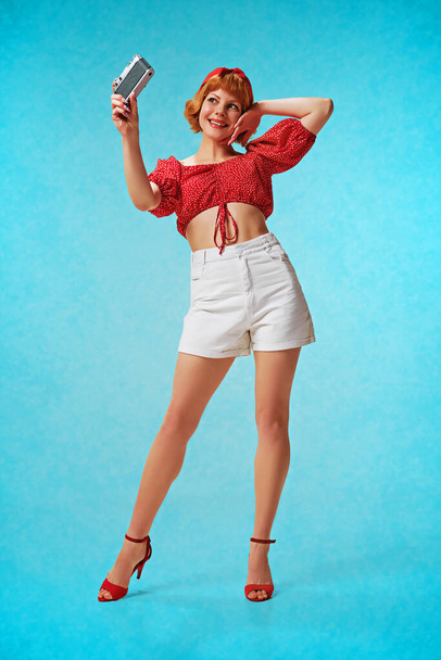 Full-length portrait of young lovely girl in attractive clothes posing, taking photo, selfie with vintage camera over blue background. Concept of retro fashion, beauty, 50s, 60s. Pin-up style - 写真・画像