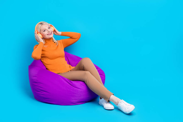 Full length φωτογραφία της καλής διάθεσης γυναίκα ντυμένος πορτοκαλί φούτερ κάθεται στα χέρια pouf αγγίζοντας ακουστικά που απομονώνονται σε μπλε φόντο χρώμα. - Φωτογραφία, εικόνα