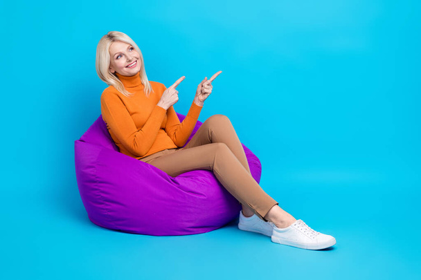 Full length photo of cheerful woman orange sweatshirt sitting on pouf look directing at sale empty space isolated on blue color background. - Фото, зображення