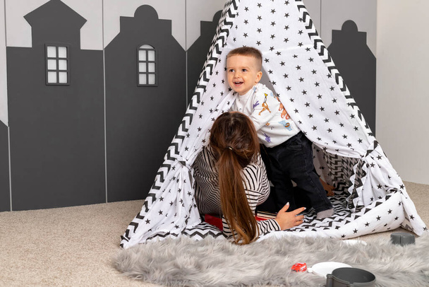 Μια μητέρα και ο γιος της παίζουν κρυφτό μέσα σε μια μικρή σκηνή στο playroom. Μωρό και νταντά περνούν χρόνο μαζί.. - Φωτογραφία, εικόνα