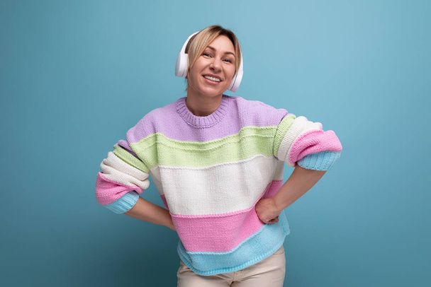niedliche attraktive blonde Frau in einem gestreiften Pullover mit großen drahtlosen Kopfhörern genießt einen Podcast auf blauem Hintergrund. - Foto, Bild