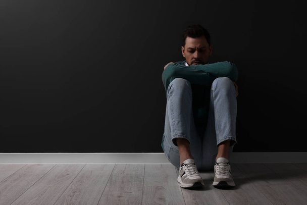 Upset man sitting on floor near black wall. Space for text - Foto, afbeelding