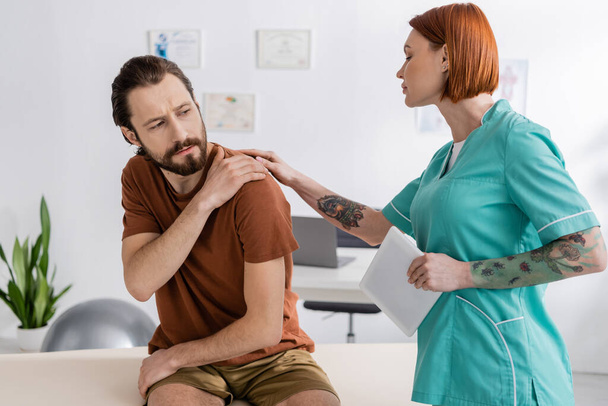 φυσιοθεραπευτής με τατουάζ με ψηφιακό tablet που αγγίζει τραυματισμένο ώμο γενειοφόρου άνδρα κατά τη διάρκεια του ραντεβού στο κέντρο αποτοξίνωσης - Φωτογραφία, εικόνα