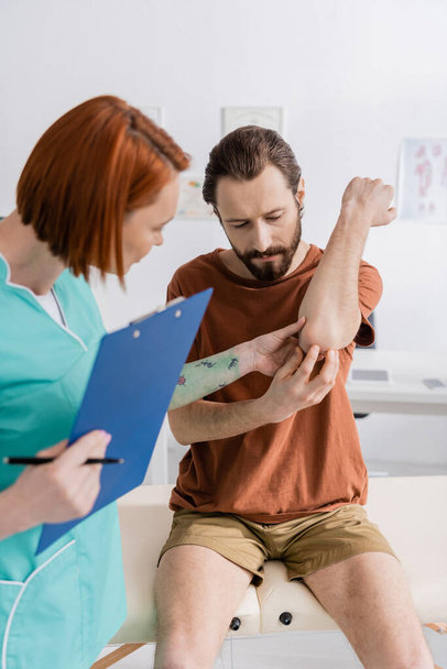 physiotherapist with digital tablet examining painful elbow of bearded man in hospital - Photo, Image