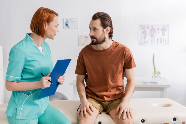 redhead rehabilitation therapist with clipboard talking to bearded man during appointment in consulting room - Photo, Image