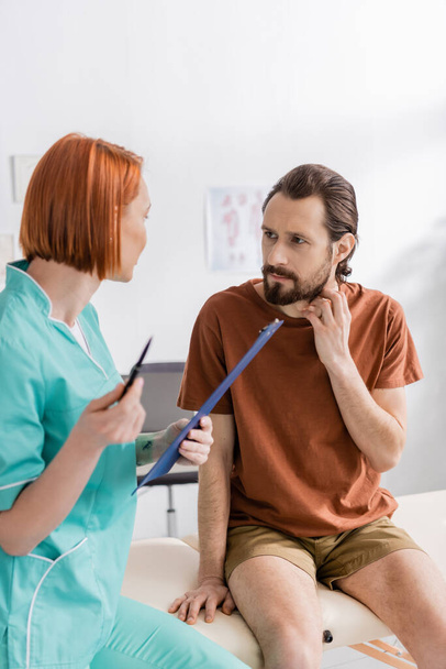roodharige fysiotherapeut met klembord in gesprek met een man met baard die de gewonde nek aanraakt in de spreekkamer - Foto, afbeelding