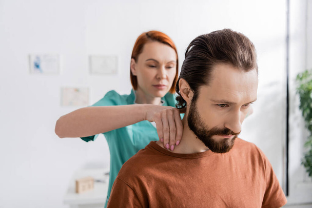 blurred osteopath touching painful neck of bearded man during examination in consulting room - Photo, image