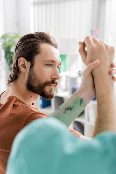 бородатий чоловік з травмованою рукою біля розмитого фізіотерапевта в реабілітаційному центрі
 - Фото, зображення