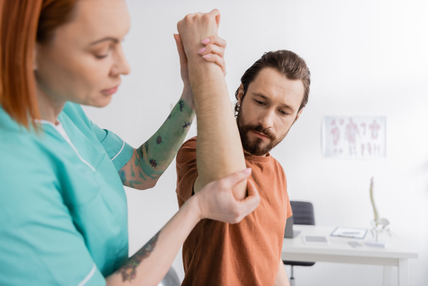 tattooed physiotherapist flexing arm of bearded man while examining injured elbow in consulting room - Foto, Imagen