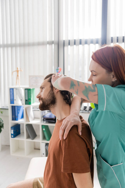 side view of tattooed chiropractor examining painful neck of bearded man in consulting room - Photo, Image