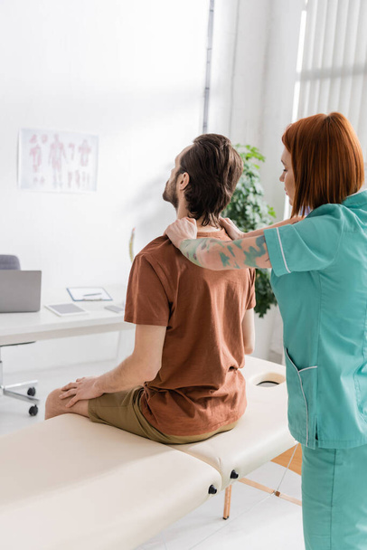 redhead osteopath touching painful back of man sitting on massage table in consulting room - 写真・画像