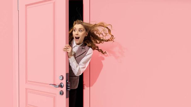 Young pretty girl peeking out open pink door and looking with positive excitement, astonishment. Surprised look. Good news, sales. Concept of emotions, facial expression, lifestyle - Φωτογραφία, εικόνα
