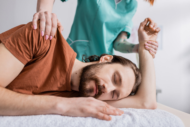 ruční terapeut masírující poraněné rameno vousatého muže se zavřenýma očima v rehabilitačním centru - Fotografie, Obrázek