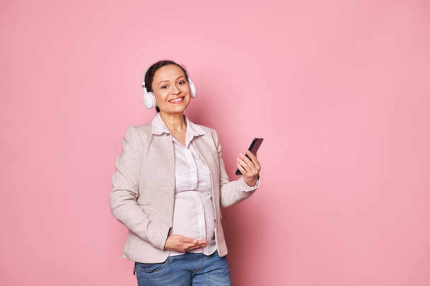 Красивая очаровательная беременная многонациональная женщина в беспроводных наушниках, наслаждается успокаивающей музыкой, улыбаясь позируя со смартфоном, прокручивая плейлист с прохладными треками, изолированные на розовом фоне - Фото, изображение