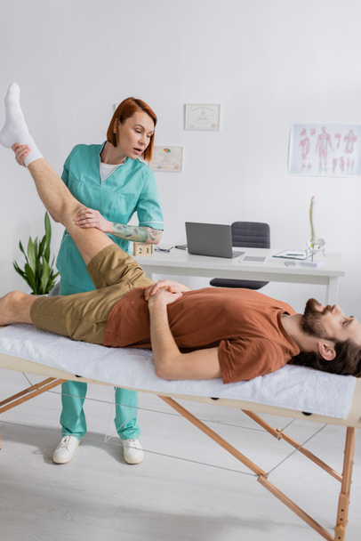 physiotherapist stretching leg of injured bearded man lying on massage table in consulting room - Valokuva, kuva