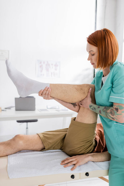 πλαϊνή όψη της κοκκινομάλλας φυσιοθεραπεύτρια που εργάζεται με τραυματισμένο πόδι ασθενούς κατά τη διάρκεια θεραπείας αποκατάστασης στην κλινική - Φωτογραφία, εικόνα