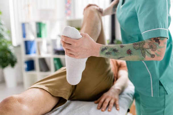cropped άποψη του τατουάζ χειροπρακτικός κάμψη πόδι του ασθενούς κατά τη διάρκεια της θεραπείας στο κέντρο αποκατάστασης - Φωτογραφία, εικόνα