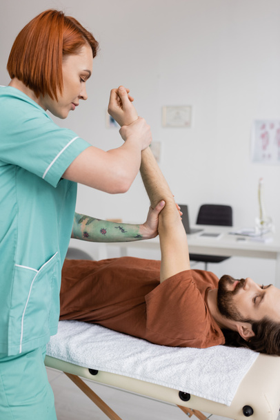 roodharige fysiotherapeut stretching pijnlijke arm van bebaarde man liggend op massagetafel in revalidatiecentrum - Foto, afbeelding