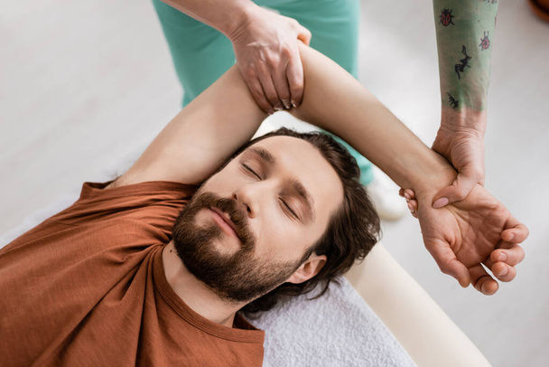 vue de dessus de l'homme barbu avec les yeux fermés près du physiothérapeute faisant massage des bras pendant la réadaptation à la clinique - Photo, image