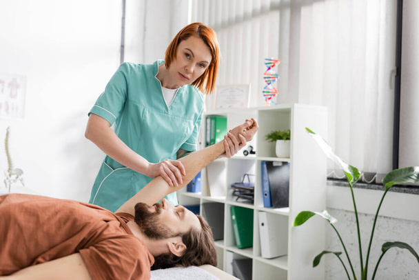 redhead physiotherapist looking at camera while working with injured arm of patient in rehab center - Fotografie, Obrázek