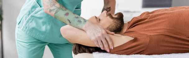 terapeuta manual tatuado haciendo masaje de alivio del dolor al hombre lesionado en el centro de rehabilitación, pancarta - Foto, imagen